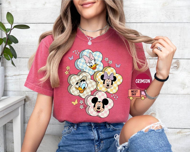 May include: A red t-shirt featuring a floral design with cartoon characters. The flowers have checkered patterns and feature Mickey Mouse, Minnie Mouse, Donald Duck, and Daisy Duck. Small butterflies and flowers are also included in the design.