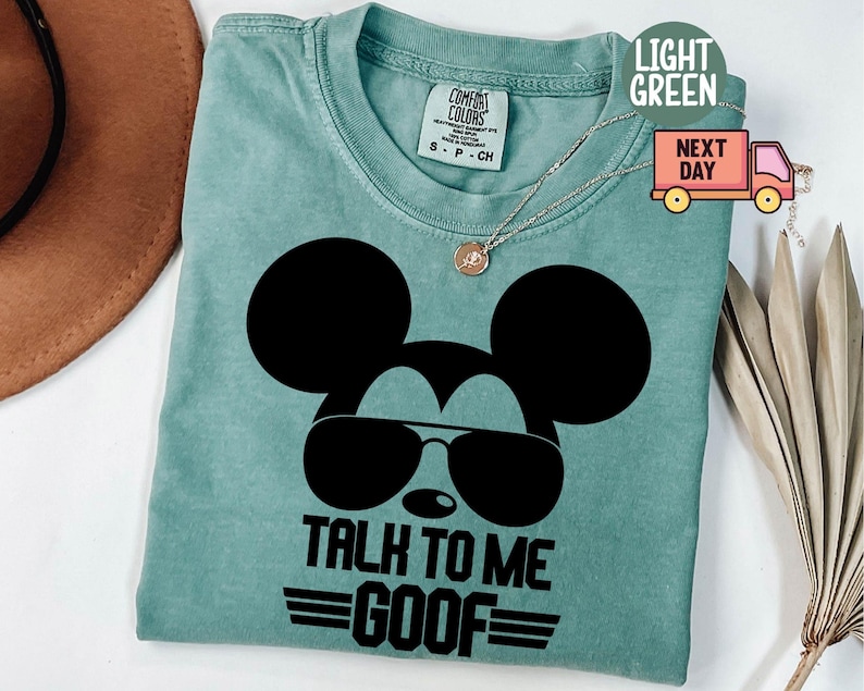 May include: A light green t-shirt featuring a black graphic of a mouse silhouette wearing sunglasses. Below the graphic, the text reads "TALK TO ME GOOF" with horizontal lines. The shirt has a Comfort Colors brand label at the neck and a thin silver chain.