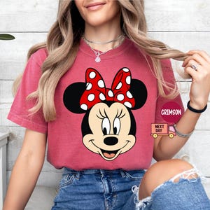 Może przedstawiać: Czerwony T-shirt z grafiką Minnie Mouse w czerwono-białej kokardce w kropki. Koszulka wykonana jest z miękkiego, wygodnego materiału.