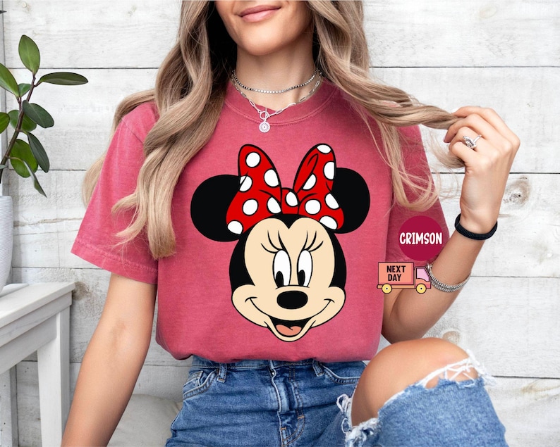 Może przedstawiać: Czerwony T-shirt z grafiką Minnie Mouse w czerwono-białej kokardce w kropki. Koszulka wykonana jest z miękkiego, wygodnego materiału.