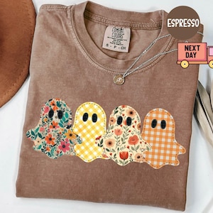 May include: Espresso-coloured t-shirt with four ghost appliqués. The ghosts have floral and gingham patterns and black dot eyes. A silver necklace with a pendant is visible. The shirt is a Comfort Colors brand.