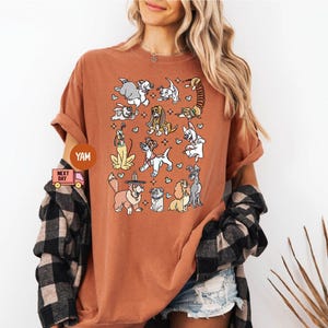 May include: A rust-colored t-shirt featuring a whimsical illustration of various dog breeds. The design includes cartoon-style dogs in different poses, with hearts and stars. The shirt has short sleeves and a relaxed fit.