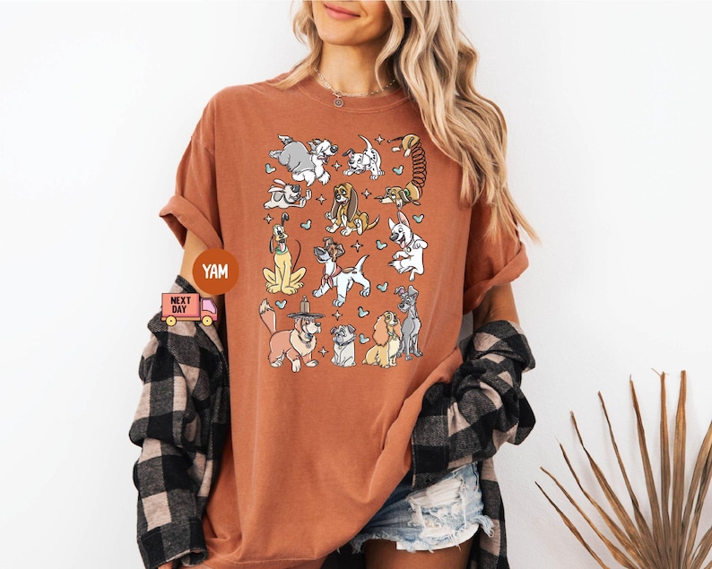 May include: A rust-colored t-shirt featuring a whimsical illustration of various dog breeds. The design includes cartoon-style dogs in different poses, with hearts and stars. The shirt has short sleeves and a relaxed fit.