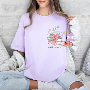 May include: A light purple t-shirt featuring a stack of books with a pink bow, stars, and the text "MRS. JONES". The design includes an open book and a delivery truck.