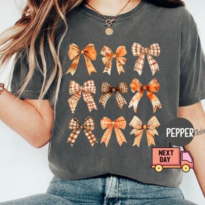 May include: A gray t-shirt with a pattern of nine brown and orange bows. The bows are arranged in three rows of three. The shirt has a distressed look and is made of a soft, comfortable fabric.