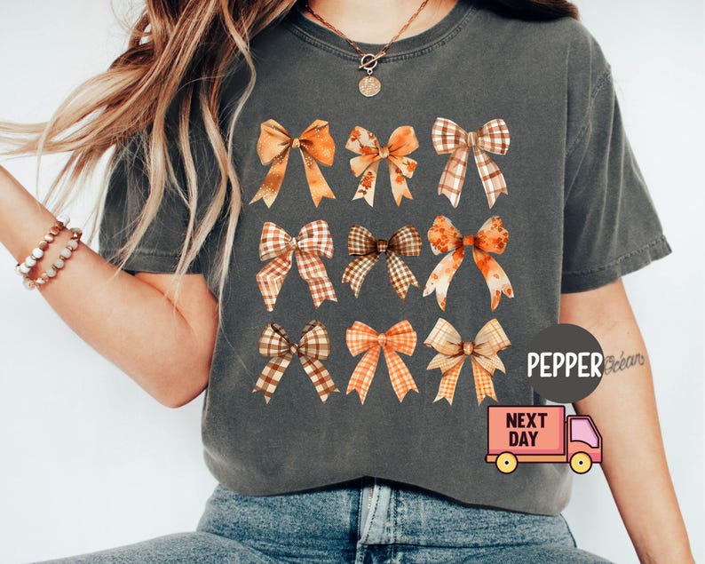 May include: A gray t-shirt with a pattern of nine brown and orange bows. The bows are arranged in three rows of three. The shirt has a distressed look and is made of a soft, comfortable fabric.