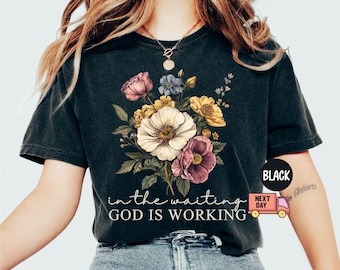 May include: Black t-shirt featuring a floral bouquet in shades of pink, yellow, and white. The text "in the waiting GOD IS WORKING" is printed below the flowers. The shirt has a vintage, washed look.