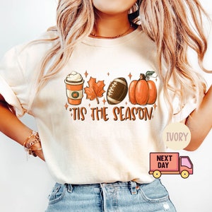 May include: A white t-shirt with a graphic design featuring a pumpkin, a football, a maple leaf, and a cup of coffee. The text "'Tis the season" is printed below the graphic. The shirt is available in ivory.
