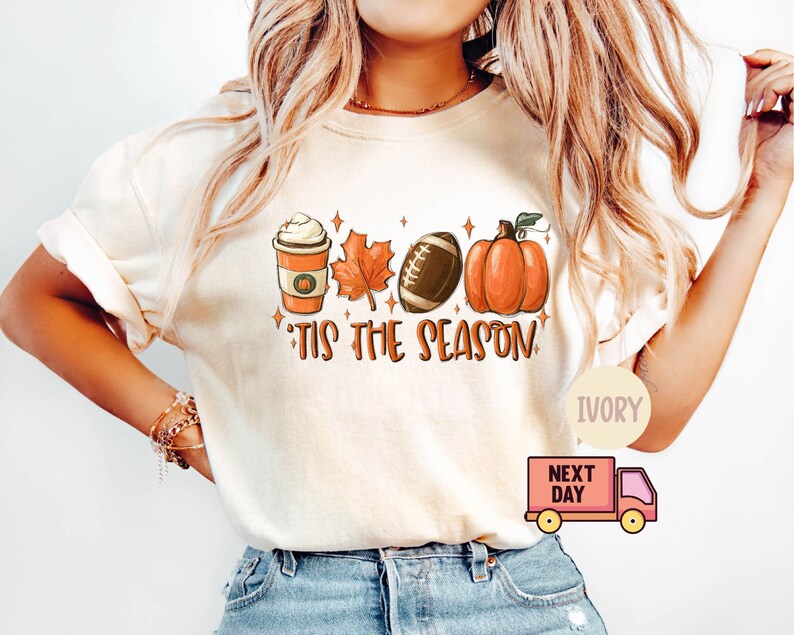 May include: A white t-shirt with a graphic design featuring a pumpkin, a football, a maple leaf, and a cup of coffee. The text "'Tis the season" is printed below the graphic. The shirt is available in ivory.