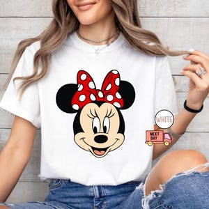 Może przedstawiać: Biała koszulka z nadrukiem czerwonej i białej Minnie Mouse. Nadruk przedstawia twarz Minnie Mouse z jej charakterystyczną czerwoną kokardką w białe kropki.