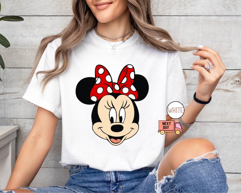 Może przedstawiać: Biała koszulka z nadrukiem czerwonej i białej Minnie Mouse. Nadruk przedstawia twarz Minnie Mouse z jej charakterystyczną czerwoną kokardką w białe kropki.