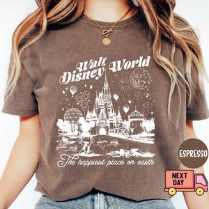May include: Espresso-colored t-shirt featuring a white graphic of Cinderella's castle, Mickey Mouse, and the text "Walt Disney World" and "The happiest place on earth." The shirt has a vintage, distressed look.