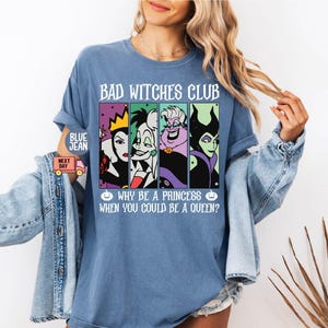 May include: A blue t-shirt with the text "BAD WITCHES CLUB" and the phrase "WHY BE A PRINCESS WHEN YOU COULD BE A QUEEN?" The shirt features four cartoon images of Disney villains. The shirt is a light blue color.