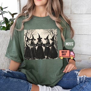 May include: Green t-shirt with a graphic print of four witches wearing black pointed hats and robes. The text "Girls Will Be Girls" is printed on the shirt.