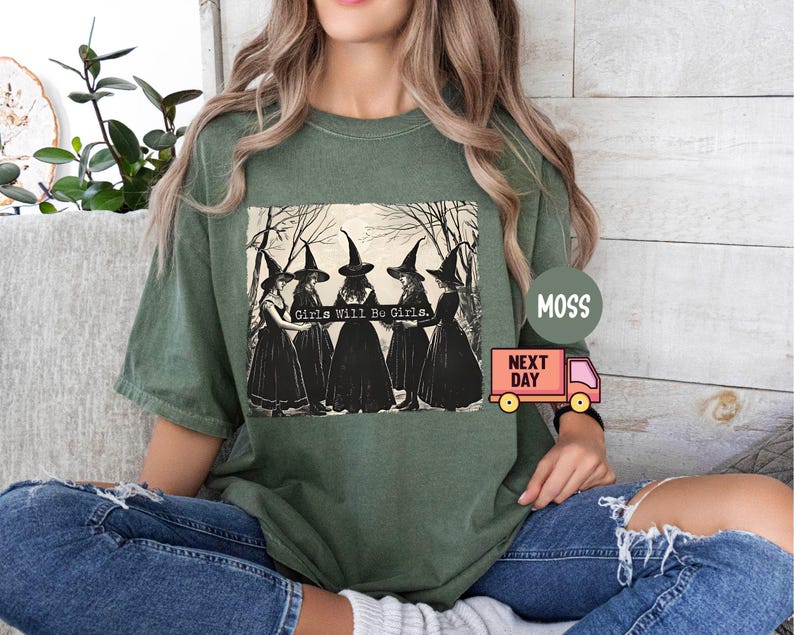 May include: Green t-shirt with a graphic print of four witches wearing black pointed hats and robes. The text "Girls Will Be Girls" is printed on the shirt.