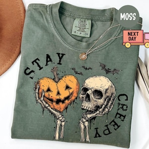 May include: A moss green crew neck t-shirt featuring a graphic of two skeletal hands holding a heart-shaped jack-o'-lantern and a human skull. The text "STAY CREEPY" is printed in black lettering around the graphic, accompanied by small bat illustrations.