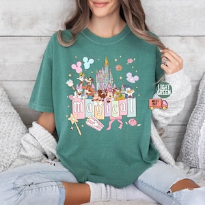 May include: Light green t-shirt featuring a colorful graphic of a castle, cartoon characters, and the word "magical" in pink letters. The design includes Mickey Mouse, Minnie Mouse, Donald Duck, and other characters. The shirt has a relaxed fit.