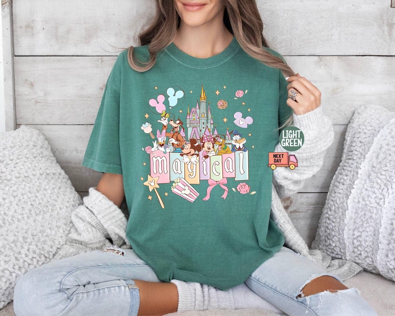 May include: Light green t-shirt featuring a colorful graphic of a castle, cartoon characters, and the word "magical" in pink letters. The design includes Mickey Mouse, Minnie Mouse, Donald Duck, and other characters. The shirt has a relaxed fit.