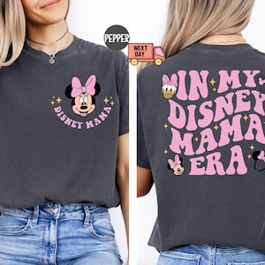 May include: Dark gray t-shirt with a pink Minnie Mouse graphic and the words "Disney Mama" in a circular design. The back of the shirt says "In My Disney Mama Era" in pink, with additional Disney character graphics.