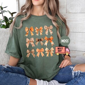 May include: A green t-shirt with a pattern of orange and brown bows. The shirt has a crew neck and short sleeves. The text "Moss Next Day" is printed on the shirt.