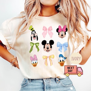 May include: Ivory-colored t-shirt featuring cartoon character faces and bows. Characters include Mickey Mouse, Minnie Mouse, Donald Duck, Goofy, and Daisy Duck. The shirt also has a pink delivery truck graphic and the word "IVORY".