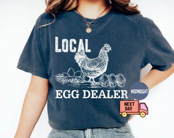 May include: Navy blue t-shirt with the words "LOCAL EGG DEALER" in white block letters. A detailed illustration of a hen, eggs, and an egg carton is printed on the shirt. The shirt is a comfortable, casual style.