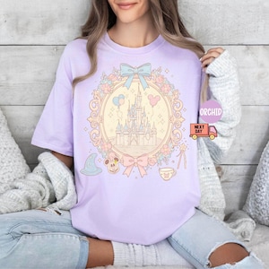 K&ouml;nnte beinhalten: Ein lavendelfarbenes T-Shirt mit einer pastellrosa und blauen Illustration von Schloss Aschenputtel, umgeben von Blumen und einer Schleife. Das Schloss ist von einem goldenen Rahmen mit hellgelbem Hintergrund umgeben. Der Text "Orchid Next Day" ist in der oberen rechten Ecke des Bildes sichtbar.