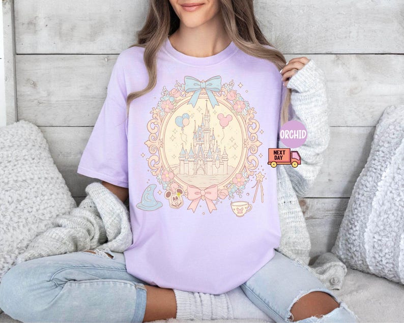 K&ouml;nnte beinhalten: Ein lavendelfarbenes T-Shirt mit einer pastellrosa und blauen Illustration von Schloss Aschenputtel, umgeben von Blumen und einer Schleife. Das Schloss ist von einem goldenen Rahmen mit hellgelbem Hintergrund umgeben. Der Text "Orchid Next Day" ist in der oberen rechten Ecke des Bildes sichtbar.