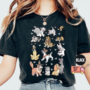 May include: Black t-shirt featuring a colorful graphic of various cartoon dogs. The design includes dogs of different breeds and poses, with small stars and butterflies. The shirt has a classic crew neck and short sleeves.