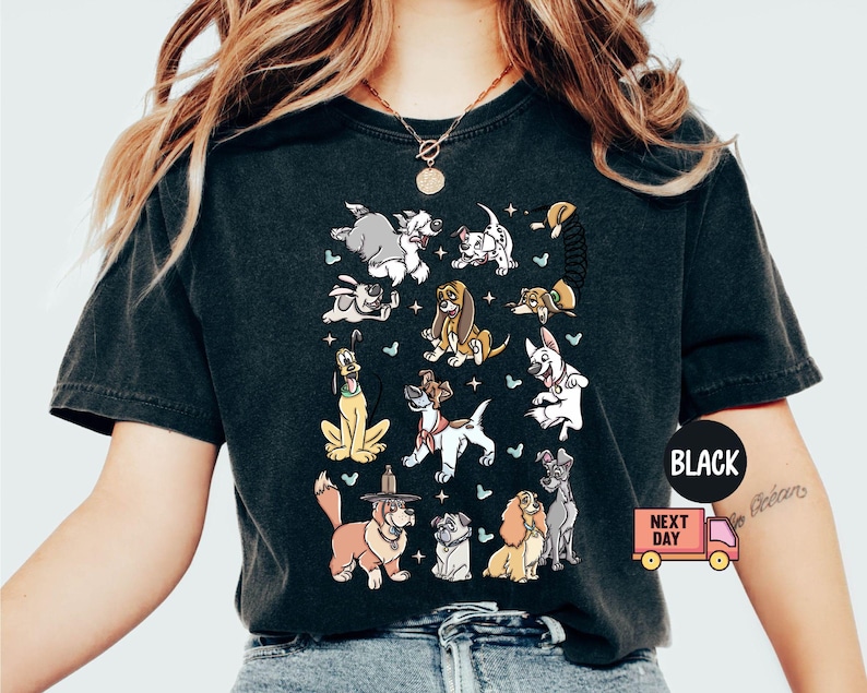 May include: Black t-shirt featuring a colorful graphic of various cartoon dogs. The design includes dogs of different breeds and poses, with small stars and butterflies. The shirt has a classic crew neck and short sleeves.