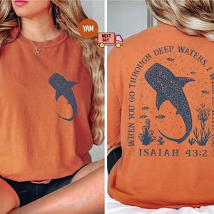 Op de afbeelding: Oranje T-shirt met een walvishaai-afbeelding op de voorkant en de tekst "WHEN YOU GO THROUGH DEEP WATERS I WILL BE WITH YOU ISAIAH 43:2" op de achterkant.