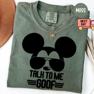 May include: A moss green short-sleeve t-shirt featuring a black graphic of a mouse silhouette wearing aviator sunglasses. Below the graphic, the text reads "TALK TO ME GOOF" with horizontal lines above and below the word "GOOF". The shirt has a crew neckline and a size small tag.