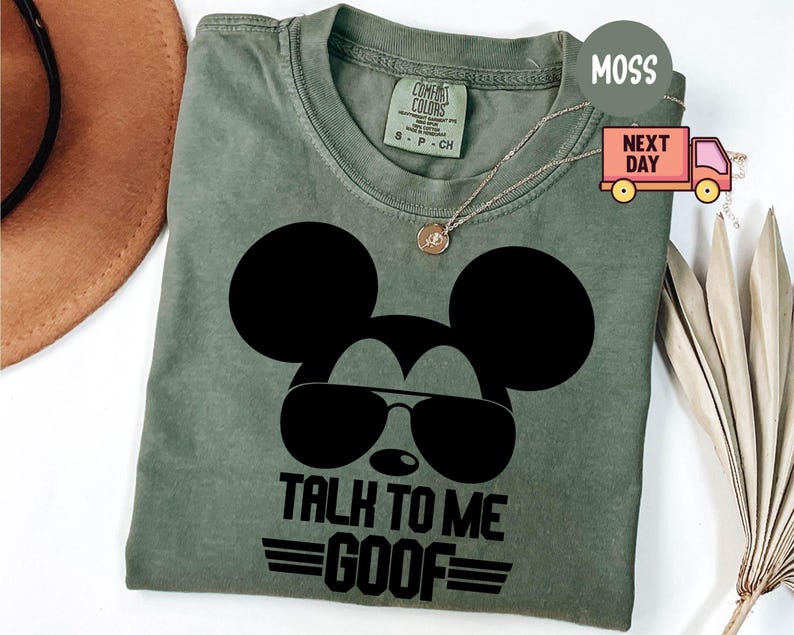 May include: A moss green short-sleeve t-shirt featuring a black graphic of a mouse silhouette wearing aviator sunglasses. Below the graphic, the text reads "TALK TO ME GOOF" with horizontal lines above and below the word "GOOF". The shirt has a crew neckline and a size small tag.