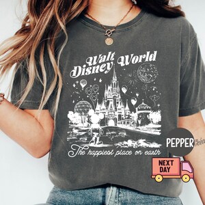 May include: Dark gray t-shirt featuring a white graphic of the Walt Disney World castle, Mickey Mouse, and fireworks. The text "Walt Disney World" and "The happiest place on earth" are also displayed. The shirt has a crew neck.
