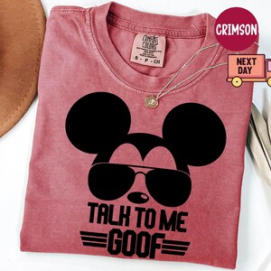 May include: A crimson short-sleeved t-shirt featuring a black graphic of a mouse silhouette wearing sunglasses. Below the graphic, the text reads TALK TO ME GOOF in bold black capital letters. The shirt has a crew neckline and a Comfort Colors label.