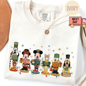 Puede incluir: Camiseta Comfort Colors color marfil con un diseño festivo de personajes de dibujos animados vestidos de cascanueces. Los personajes están dispuestos en fila, con detalles coloridos y luces navideñas. La camiseta tiene cuello redondo.