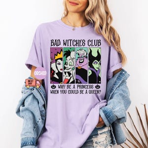 May include: A light purple t-shirt with the text "BAD WITCHES CLUB" and the phrase "WHY BE A PRINCESS WHEN YOU COULD BE A QUEEN?". The shirt features four cartoon images of Disney villains. The shirt is worn by a person.