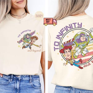 May include: A cream-colored t-shirt with a vintage-style graphic of Woody and Buzz Lightyear from Toy Story. The graphic features the text "To Infinity and Beyond" and a retro-style design with stars and stripes.