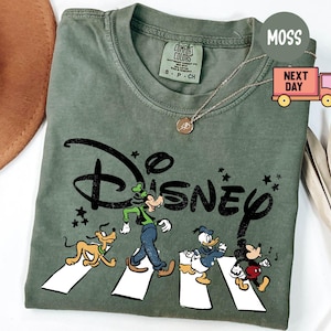 K&ouml;nnte beinhalten: Moosgr&uuml;nes T-Shirt mit einem Disney-Motiv. Das Design zeigt Goofy, Pluto, Donald Duck und Mickey Mouse beim &Uuml;berqueren einer Stra&szlig;e. Das Wort "Disney" ist in einer schwarzen, stilisierten Schriftart gehalten. Das T-Shirt hat einen Rundhalsausschnitt und kurze &Auml;rmel.