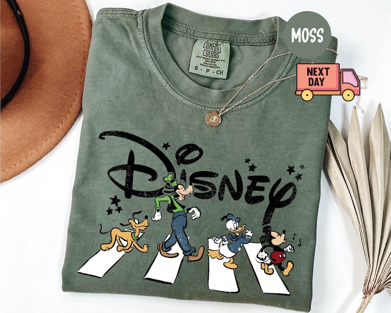 K&ouml;nnte beinhalten: Moosgr&uuml;nes T-Shirt mit einem Disney-Motiv. Das Design zeigt Goofy, Pluto, Donald Duck und Mickey Mouse beim &Uuml;berqueren einer Stra&szlig;e. Das Wort "Disney" ist in einer schwarzen, stilisierten Schriftart gehalten. Das T-Shirt hat einen Rundhalsausschnitt und kurze &Auml;rmel.