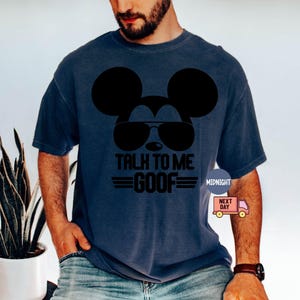 May include: A dark blue short-sleeved t-shirt featuring a black graphic of a mouse silhouette wearing sunglasses. Below the graphic, the text reads TALK TO ME GOOF in bold black letters, flanked by two horizontal lines on each side of the word GOOF.