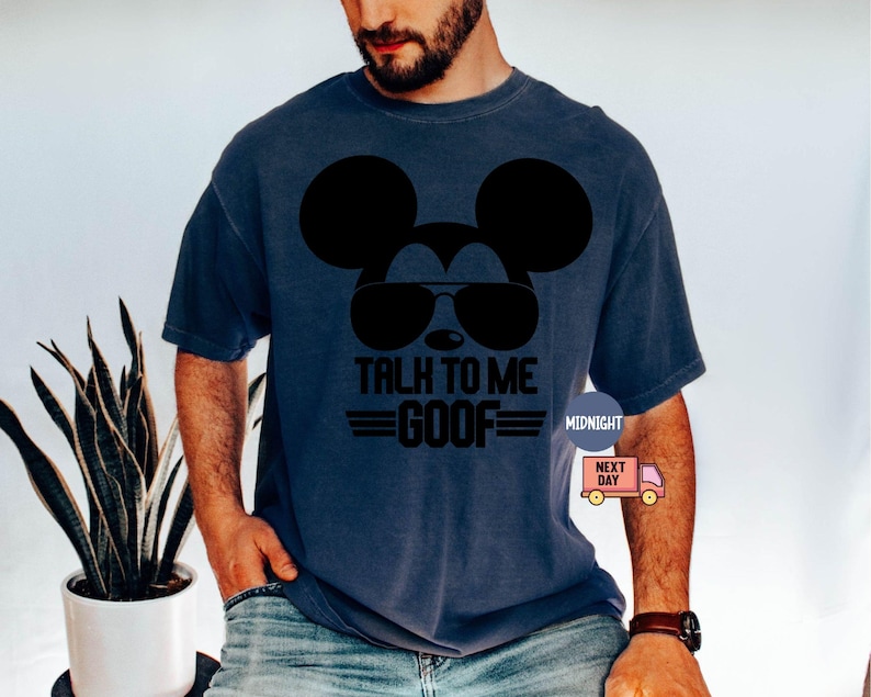 May include: A dark blue short-sleeved t-shirt featuring a black graphic of a mouse silhouette wearing sunglasses. Below the graphic, the text reads TALK TO ME GOOF in bold black letters, flanked by two horizontal lines on each side of the word GOOF.
