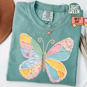 以下が含まれることがあります： パッチワークの蝶のデザインが施された、明るい緑色のTシャツ。蝶の羽は、ピンク、黄色、青など、さまざまな色と模様があります。シャツは丸いネックラインと半袖です。小さなペンダントが付いたシルバーのネックレスがシャツの上にあります。