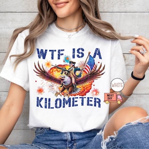 May include: White t-shirt with a graphic featuring an eagle carrying a person with a gun and an American flag. The text on the shirt reads "WTF IS A KILOMETER."