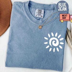 May include: A blue jean Comfort Colors t-shirt with a white sun graphic. The shirt has a crew neck and short sleeves. A gold necklace with a small pendant is on the shirt.