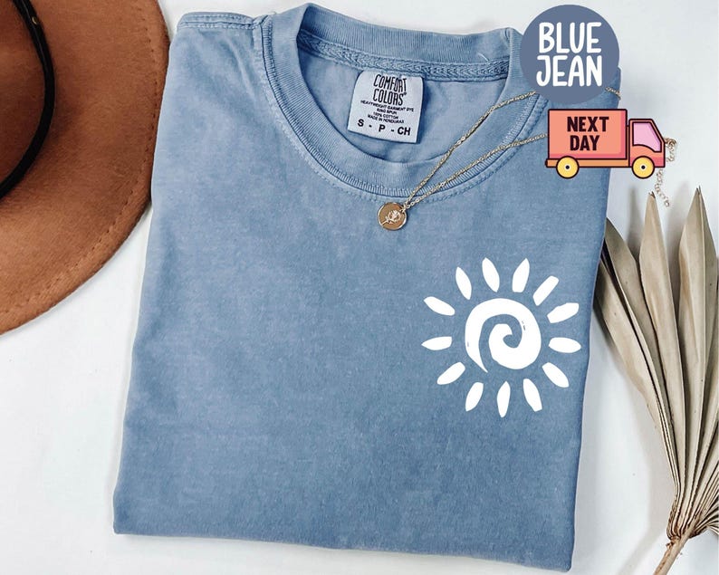 May include: A blue jean Comfort Colors t-shirt with a white sun graphic. The shirt has a crew neck and short sleeves. A gold necklace with a small pendant is on the shirt.