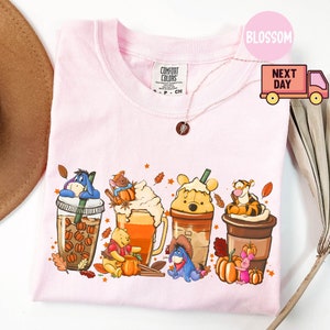 May include: Light pink t-shirt featuring a colorful graphic of Winnie the Pooh characters in fall-themed coffee drinks. The shirt has a crew neck and short sleeves. The brand name "Comfort Colors" is visible on the shirt.