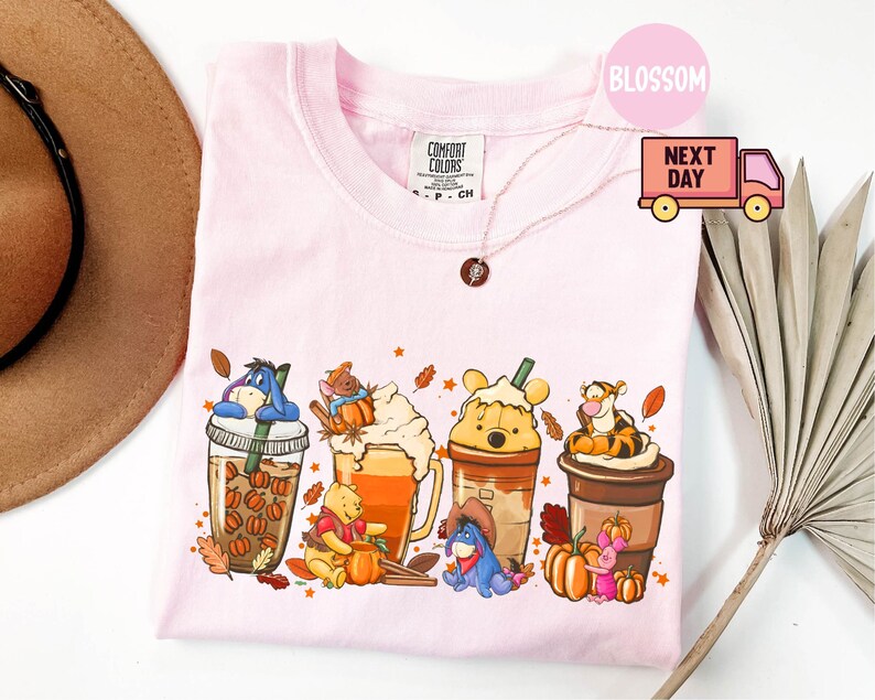 May include: Light pink t-shirt featuring a colorful graphic of Winnie the Pooh characters in fall-themed coffee drinks. The shirt has a crew neck and short sleeves. The brand name "Comfort Colors" is visible on the shirt.