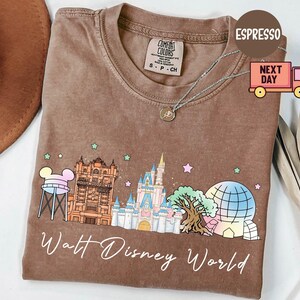 May include: Espresso-colored t-shirt featuring a colorful graphic of Walt Disney World landmarks. The design includes Cinderella's castle, the Hollywood Tower Hotel, and Spaceship Earth. The text "Walt Disney World" is written in a cursive font.