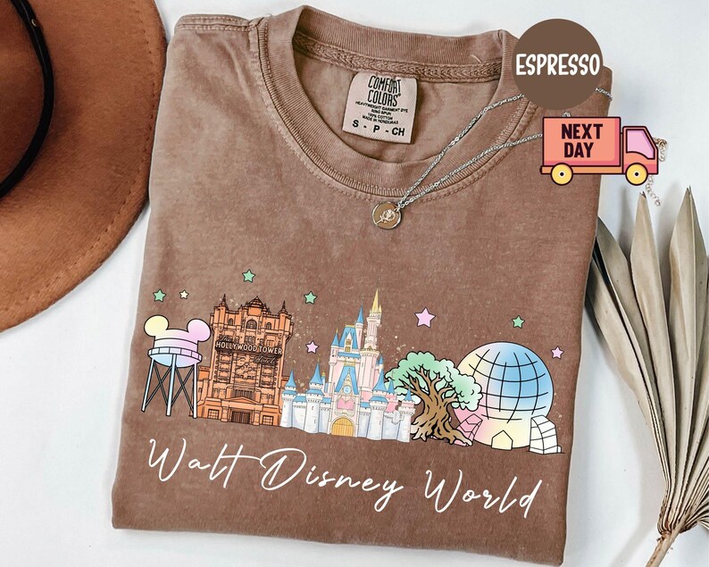 May include: Espresso-colored t-shirt featuring a colorful graphic of Walt Disney World landmarks. The design includes Cinderella's castle, the Hollywood Tower Hotel, and Spaceship Earth. The text "Walt Disney World" is written in a cursive font.